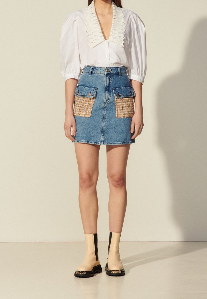 Femme portant un chemisier blanc à col en dentelle, une jupe en jean avec des poches avant à carreaux et en denim, et des bottines beiges à semelles noires.
