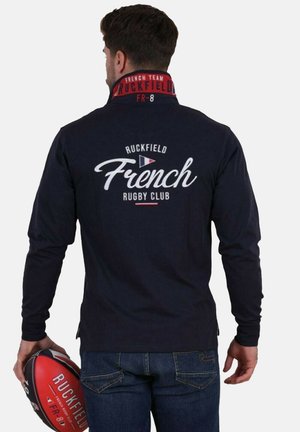 Homme portant une chemise à manches longues bleu marine avec le texte "Ruckfield French Rugby Club" dans le dos, tenant un ballon de rugby rouge portant l'inscription "Ruckfield FR-8."