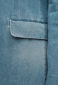 Tissu en denim bleu clair avec une poche à rabat horizontale et des détails de coutures visibles sur un vêtement.