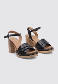 Sandalias de cuero negro con una tira ancha, tira en el tobillo para un ajuste seguro y un tacón cuadrado. La suela tiene un color marrón claro contrastante y un detalle de costura.