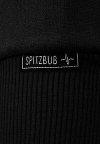 Zwarte ribstoffen met een label waarop "SPITZBUB" in witte tekst staat. Het label omvat een lijngrafiek en is geplaatst op een textuurrijke zone.