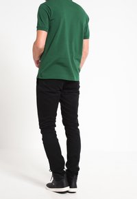 Polo en coton vert à manches courtes, associé à un pantalon slim noir et des baskets grises avec semelles blanches. Vue de dos.