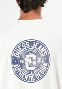 Rückseite eines weißen T-Shirts mit einem blauen, kreisförmigen Guess Jeans-Logo, das einen geflügelten Sneaker und den Text "Los Angeles CA Track & Field 1981" zeigt.