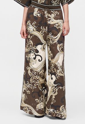 Pantalon marron à jambes larges avec motifs ondulés et tourbillonnants crème et or, taille élastique et cordon, porté par une personne ayant les mains détendues sur les côtés.