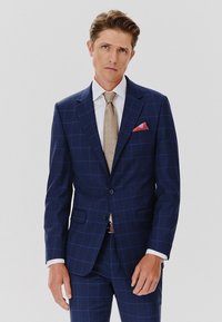 Granatowy garnitur w kratę, dopasowany krój, klapy z wykrojem oraz dwie przednie kieszenie. W zestawie z białą koszulą i beżowym krawatem.
