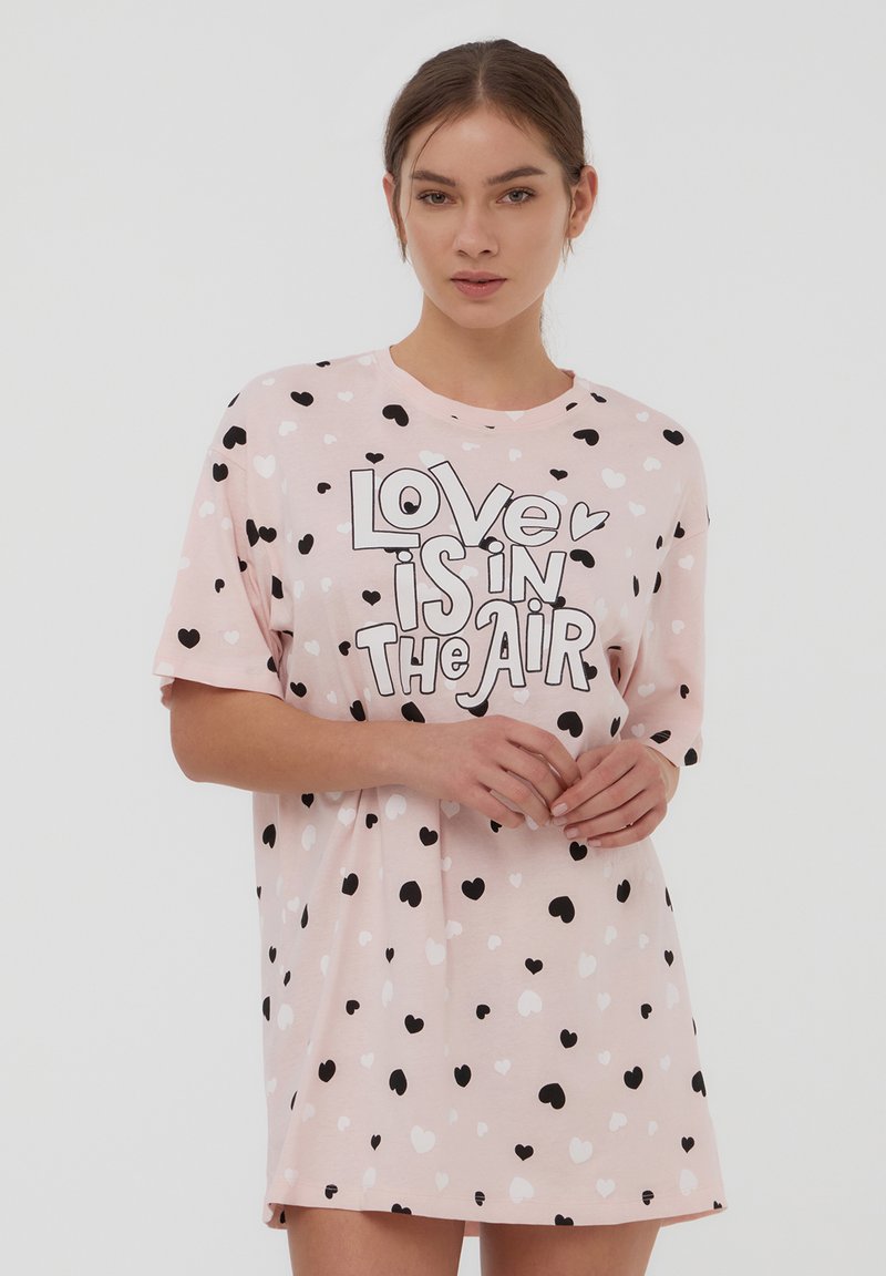 Gonna notte rosa a maniche corte con un motivo a cuori in bianco e nero. Presenta il testo "L'amore è nell'aria" in grassetto nero.
