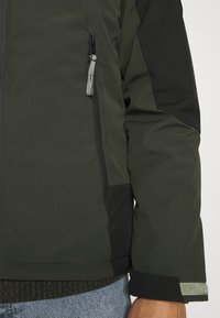 Mörkgrön jacka med svarta detaljer, har en ficka med dragkedja, en texturerad mudd och ett slätt, vattentåligt tyg.