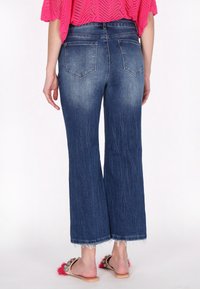 Jeans de mezclilla oscura de pierna ancha con dobladillo deshilachado, dos bolsillos traseros y una textura suave, combinados con sandalias decorativas.