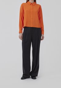 Chemise orange boutonnée avec col, associée à un pantalon noir ample. La chemise est en tissu texturé et de coupe courte.