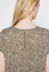 Blouse marron avec un motif de feuilles blanches, manches courtes et une fermeture éclair dorée au dos. Tissu à texture lisse.