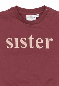 Camicia di cotone bordeaux con la parola "sorella" ricamata in crema, scollatura rotonda e bottoni a pressione su una spalla per facilitare l'indossamento.