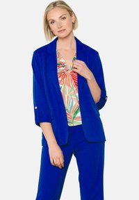 Sinine bleiser, mille varrukad on üles keeratud, koos värvilise mustriga särgiga, millel on punased ja rohelised lehed. Sile kangas, hästi istuv.