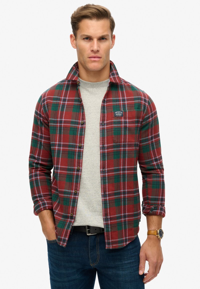 Rood en groen geblokt flanellen shirt met een patchzak, knoopsluiting en opgerolde mouwen over een grijs ronde hals t-shirt. Blauwe spijkerbroek en horloge.