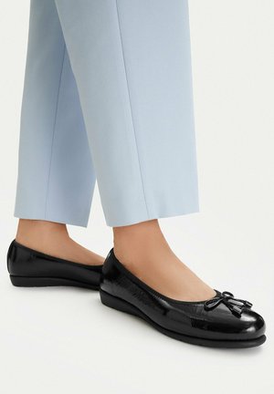 Osoba nosící světle modré kalhoty a lesklé černé baleríny s malými mašličkami na bílém pozadí.