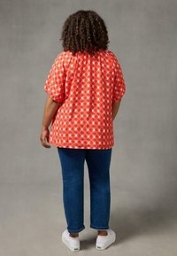 Red patterned short-sleeve shirt with geometric designs, paired with blue jeans and white sneakers. Background is a neutral gray.