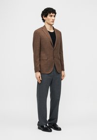 Άνδρας με καφέ σακάκι, μαύρο πουκάμισο, γκρι παντελόνι και μαύρα παπούτσια, που στέκεται μπροστά σε ένα απλό φωτεινό φόντο.