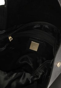 Crna kožna torba s teksturiranim vanjskim dijelom. Unutrašnjost ima glatku crnu podstavu, zippered odjeljke i zlatnu oznaku.