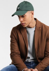 Grüne Baumwoll-Baseballkappe mit gesticktem Logo auf der Vorderseite, kombiniert mit einem grauen Sweatshirt und einem braunen Blazer. Fokus auf die Form und die Farbe der Kappe.