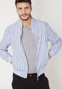 Chaqueta bomber azul claro con rayas verticales blancas, puños y cuello acanalados, que incluye cremalleras y un detalle de bolsillo frontal.