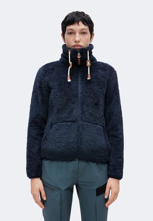 Persona con una chaqueta de forro polar de cuello alto azul oscuro con cordones y pantalones turquesa con parches negros, mirando hacia adelante sobre un fondo liso.