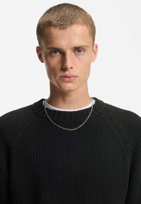 Maglione nero a maglia con colletto a coste, indossato sopra una camicia bianca, accompagnato da una semplice collana in argento.