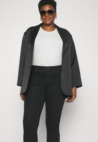 Blazer negro de satén con un corte relajado, combinado con una camiseta blanca y jeans ajustados negros. Modelo llevando gafas de sol oscuras y pendientes de aro.