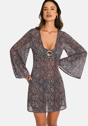 Femme portant une mini-robe transparente à manches longues avec un motif peau de serpent, un décolleté en V profond et une boucle circulaire décorative sur la poitrine.