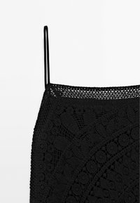 Haut en crochet noir avec fines bretelles et motifs floraux et géométriques complexes sur le tissu.
