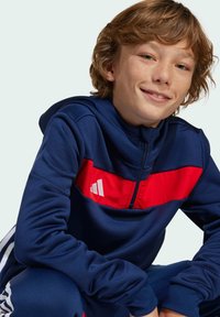 Mornarski plavi sportski dukserica s crvenom horizontalnom prugom i bijelim logotipom. Ima zip zatvarač na ovratniku i mekan, glatak materijal.