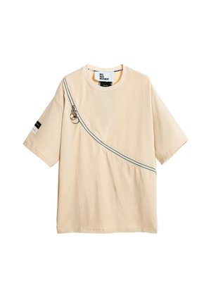 Joven con el pelo trenzado está de pie mirando hacia adelante con una camiseta beige de gran tamaño y pantalones blancos de pierna ancha con líneas curvas negras y zapatos neutros.