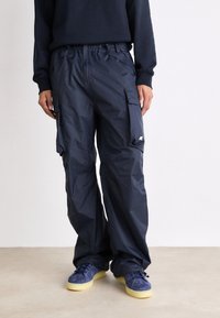 Personne portant un pantalon cargo ample bleu marine foncé avec de grandes poches latérales et des baskets bleu marine à semelles beiges, debout devant un fond uni.