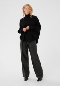 Moteris su garbanotais šviesiais plaukais, vilkinti juodą ribotos tekstūros šiltą megztinį ir laisvas juodas odines kelnes, stovi prieš paprastą foną.