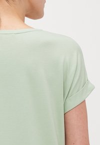Lysegrønn kortermet topp med rullede ermer, laget av myk stoff. Har en flat utringning og en jevn, subtil tekstur.