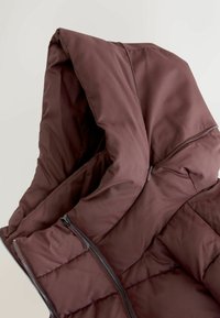 Pruun puffjakker, millel on kapuuts, tikandiga disain ja kaks külgset zipperit. Kanga pind tundub sile ja mati viimistlusega.