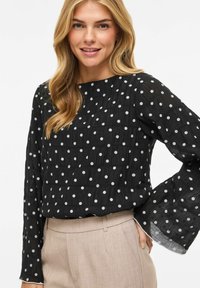 Must mustriga pluus pikkade varrukatega ja ümmarguse kaelusega. Kangas näib struktuurne ja kerge, kombineeritud beežide pükstega.