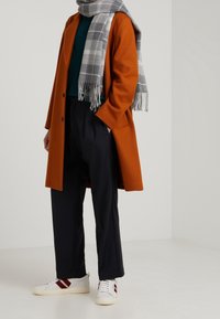 Manteau en laine orange, haut vert, écharpe écossaise grise, pantalon bleu marine et baskets blanches avec des rayures rouges. Comprend des coupes ajustées et des textures.