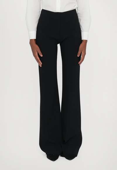 Schwarze lange Hosen mit hoher Taille, weit geschnitten, aus glattem Stoff. Kombiniert mit einem weißen Hemd mit Knöpfen.