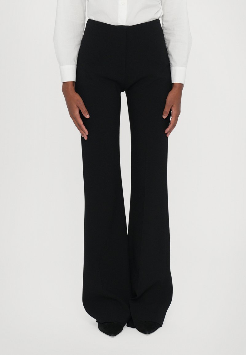Zwarte lange broek met een hoge taille, wijde pijpen en gemaakt van een gladde stof. Gepaard met een wit overhemd met knopen.