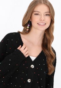 Fekete kardigán színes díszes gyöngyökkel, V-nyakkivágású, oversize fazonú, és két ezüst gombbal, csillag alakú fülbevalóval párosítva.