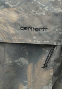 Šedá maskáčová tkanina s našitým čiernym logom "carhartt", nastaviteľná šnúrka na stiahnutie a vrecko s klopou s hladkou textúrou.