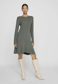 Robe tricotée verte à manches longues, texture côtelée et accents de boutons dorés sur les épaules. Associée à des bottes beiges à talons aiguilles arrivant au genou.