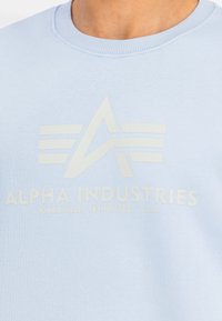 Világoskék pulóver, puha anyagból, nagy, krém színű logóval, stílusos betűtípussal és design elemekkel.