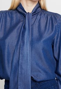 Eine Frau trägt eine blaue Denim-Bluse mit hohem Kragen und einem langen, lockeren Schlipsdetail an der Vorderseite.