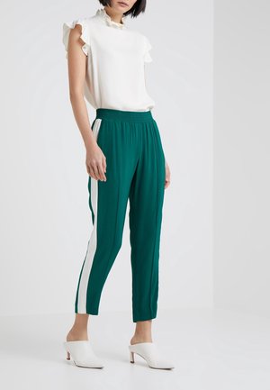 Kvinde iført hvid flæset ærmeløs top, grønne bukser med hvide sidelister og hvide mulehøjhælede sko, stående foran ensfarvet baggrund.