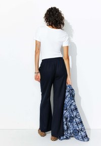 T-shirt blanc, pantalon large bleu marine décontracté, tenant une écharpe à motifs bleus et des chaussures en imprimé léopard. L'arrière-plan est blanc uni.