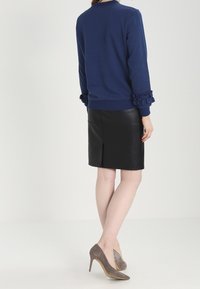 Žena v námořnickém svetru s volánovými manžetami, černé kožené sukně do kolen se zadním rozparkem a šedých semišových lodičkách na vysokém podpatku, stojí otočená zády.