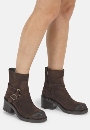 Jambes portant des bottines en daim brun foncé avec talons blocs, boucles latérales et fermetures éclair sur un fond clair.