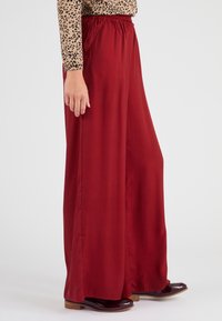 Rode wijde broek met een elastische tailleband, gemaakt van een gladde stof. De broek heeft zijdelingse naden en is gecombineerd met donker, glanzend schoeisel.
