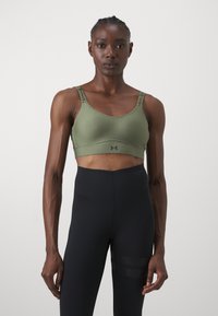 Soutien de desporto verde oliva com alças ajustáveis apresentando um logo. O design é ajustado com uma textura suave. Combinado com leggings pretas.
