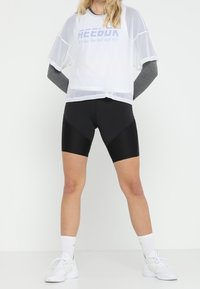 Vit mesh crop top med lila logotyp, ihop med svarta cykelshorts. Modellen bär vita sneakers och strumpor till mitten av vaden med designinslag.
